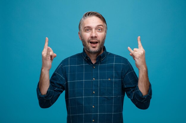 Middle age man with grey hair in dark color shirt crazy happy and excited showing rock symbols standing over blue background