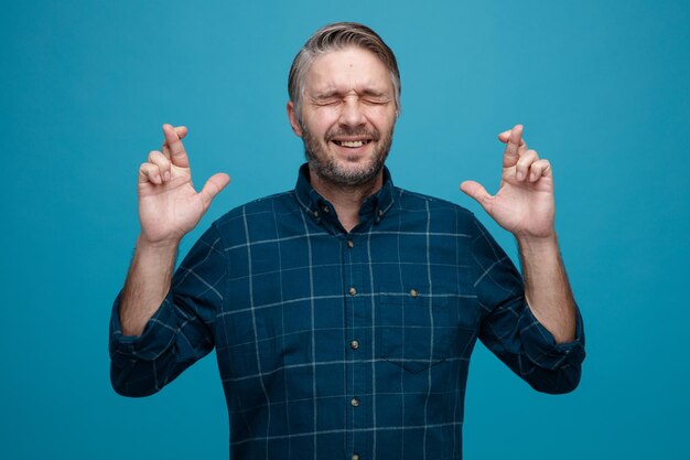 Middle age man with grey hair in dark color shirt crossing fingers making wish being excited standing over blue background