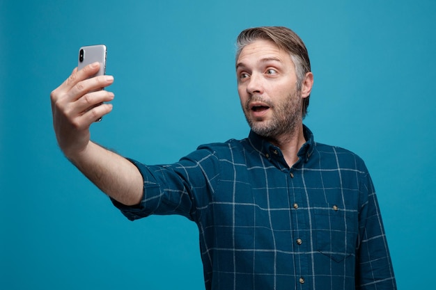 Мужчина средних лет с седыми волосами в рубашке темного цвета делает селфи с помощью смартфона и выглядит удивленным, стоя на синем фоне