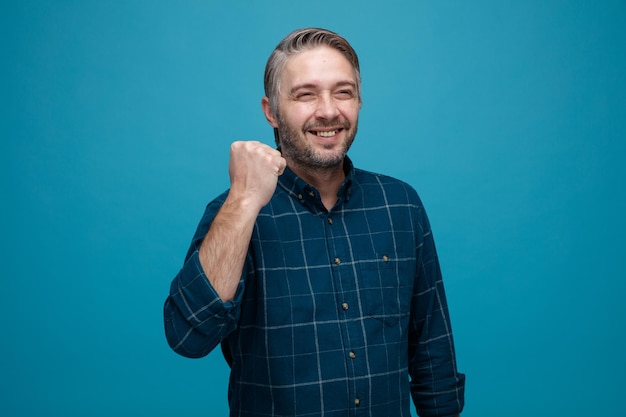 Middle age man with grey hair in dark color shirt happy and cheerful clenching fist rejoicing his success standing over blue background