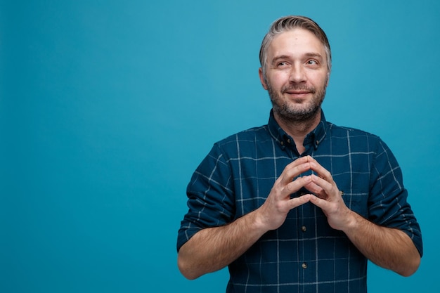 Free photo middle age man with grey hair in dark color shirt holding hands together looking aside waiting for something smiling slyly standing over blue background