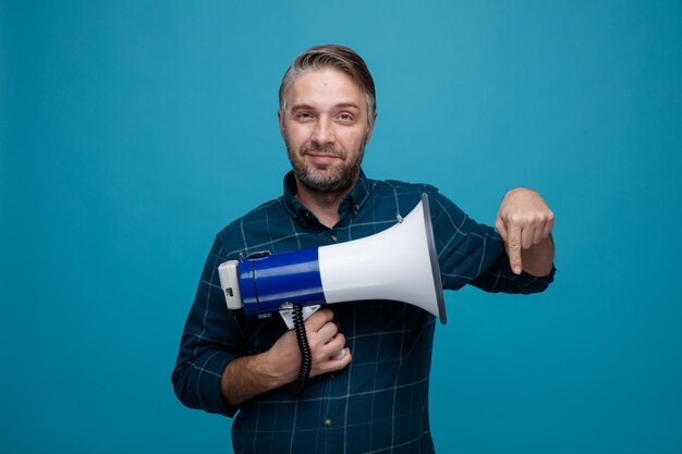 Middle age man with grey hair in dark color shirt holding megaphone looking at camera smiling pointing with index finger down standing over blue background