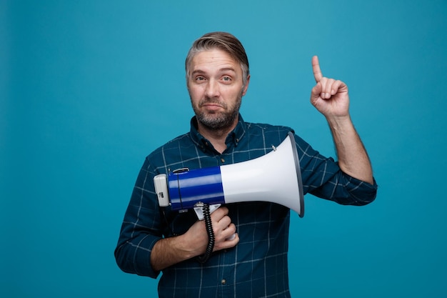 Middle age man with grey hair in dark color shirt holding megaphone looking at camera smiling pointing with index finger up standing over blue background