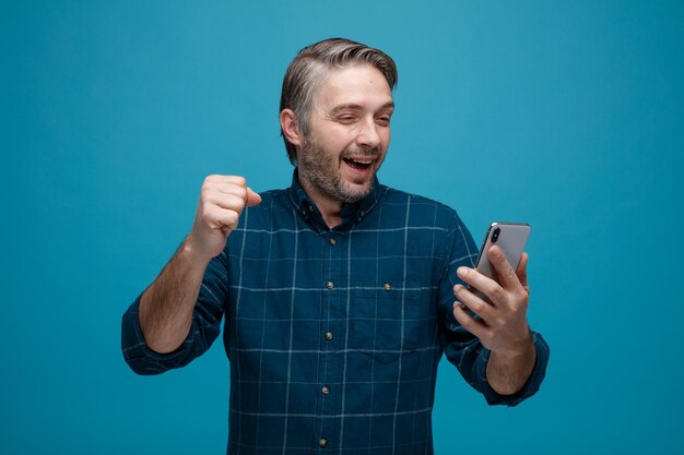 Мужчина средних лет с седыми волосами в рубашке темного цвета держит смартфон, смотрит на экран, счастливый и взволнованный, сжимая кулак, стоя на синем фоне