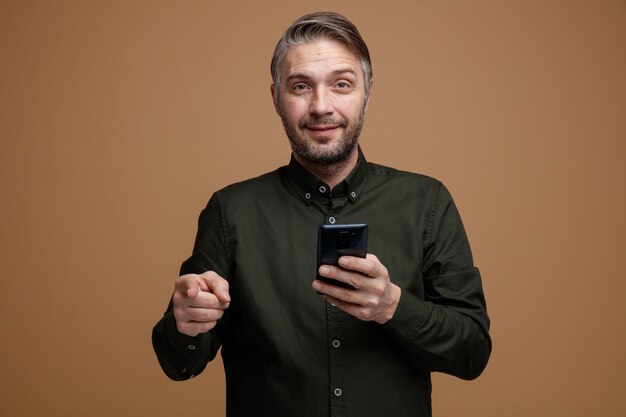 мужчина средних лет с седыми волосами в рубашке темного цвета держит смартфон, указывая указательным пальцем на камеру, улыбаясь уверенно, счастливо и позитивно, стоя на коричневом фоне