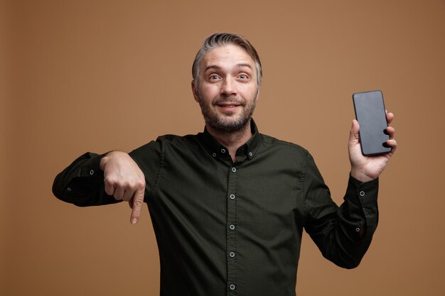 Middle age man with grey hair in dark color shirt holding smartphone pointing with index finger down looking at camera happy and positive smiling standing over brown background