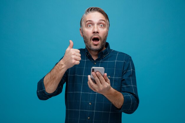 Middle age man with grey hair in dark color shirt holding smartphone showing thumb up looking at camera amazed and surprised standing over blue background