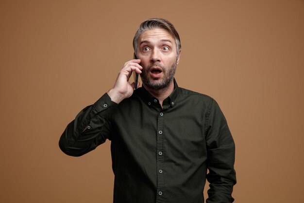 Мужчина средних лет с седыми волосами в рубашке темного цвета выглядит удивленным и удивленным, разговаривая по мобильному телефону, стоя на коричневом фоне
