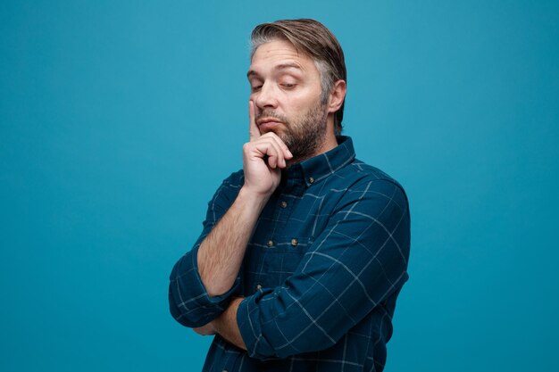 Middle age man with grey hair in dark color shirt looking aside puzzled thinking touching his cheek standing over blue background