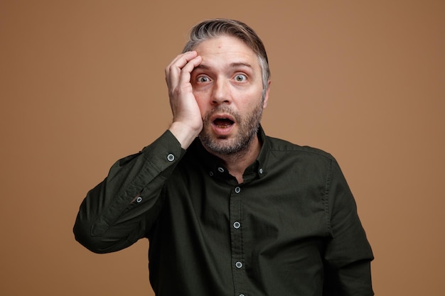 Middle age man with grey hair in dark color shirt looking at camera amazed and shocked standing over brown background
