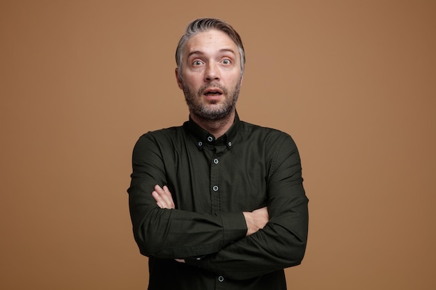 Middle age man with grey hair in dark color shirt looking at camera amazed and surprised crossing hands on his chest standing over brown background