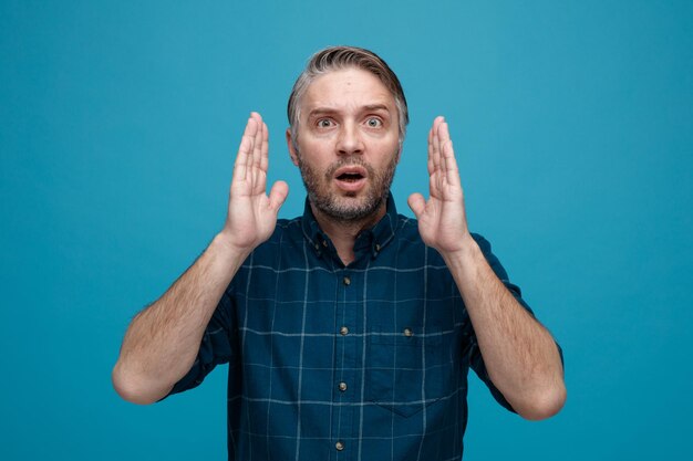 Middle age man with grey hair in dark color shirt looking at camera being amazed surprised showing size sign with hands standing over blue background