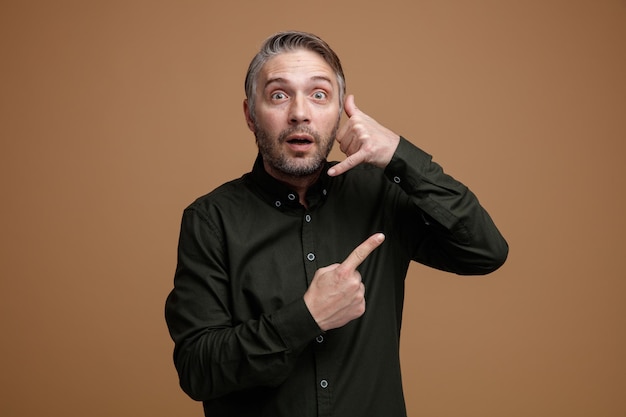 Middle age man with grey hair in dark color shirt looking at camera being surprised making call me gesture with hand pointing with index finger to the side standing over brown background