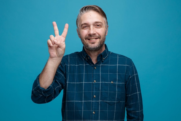 middle age man with grey hair in dark color shirt looking at camera happy and cheerful showing vsign smiling standing over blue background