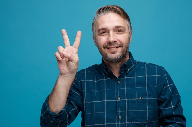 Middle age man with grey hair in dark color shirt looking at camera happy and cheerful showing vsign smiling standing over blue background