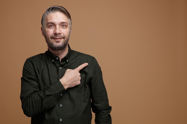 Middle age man with grey hair in dark color shirt looking at camera happy and positive pointing with index finger to the side standing over brown background