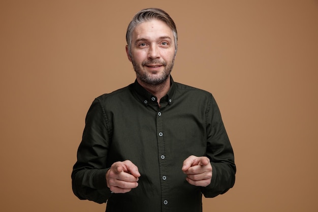 Мужчина средних лет с седыми волосами в рубашке темного цвета смотрит в камеру счастливым и позитивным, указывая указательными пальцами на камеру, стоящую на коричневом фоне