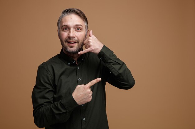 Middle age man with grey hair in dark color shirt looking at camera happy and positive smiling making call me gesture pointing with index finger to the side standing over brown background