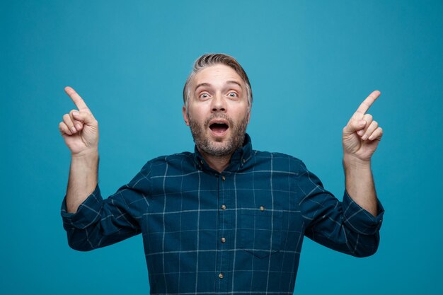 Foto gratuita uomo di mezza età con i capelli grigi in camicia di colore scuro che guarda la fotocamera felice e sorpreso che punta con il dito indice in piedi su sfondo blu