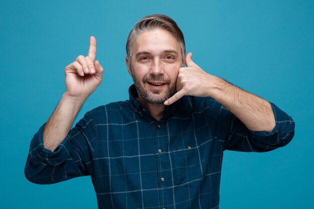 Middle age man with grey hair in dark color shirt looking at camera making call me gesture pointing with index finger up smiling with happy face standing over blue background