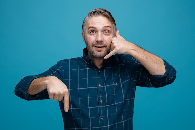 Middle age man with grey hair in dark color shirt looking at camera making call me gesture with hand pointing with index finger down smiling standing over blue background