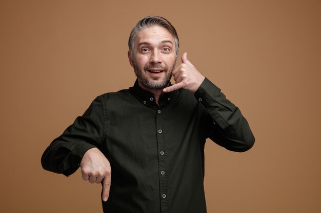 Middle age man with grey hair in dark color shirt looking at camera making call me gesture with hand pointing with index finger down smiling standing over brown background