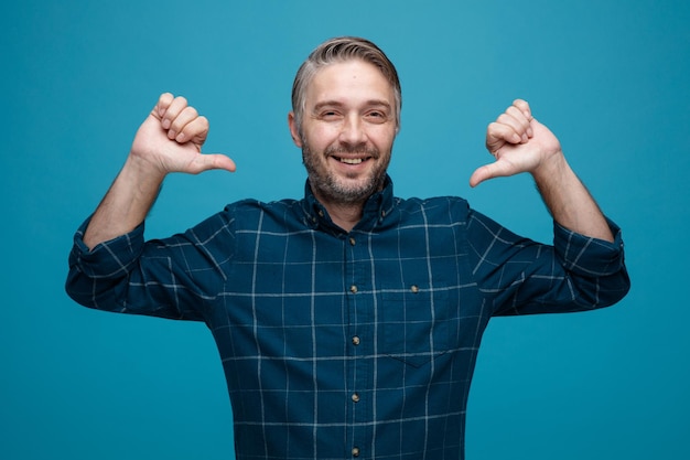 Middle age man with grey hair in dark color shirt looking at camera smiling happy and positive pointing with thumbs at himself proud and confident standing over blue background