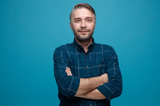 Middle age man with grey hair in dark color shirt looking at camera with confident expression with crossed hands on his chest standing over blue background