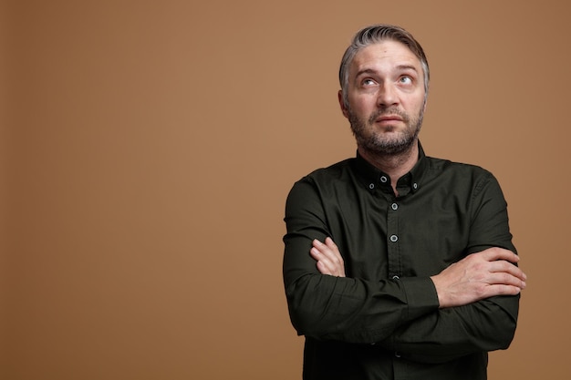 Middle age man with grey hair in dark color shirt looking up puzzled thinking crossing hands on his chest standing over brown background