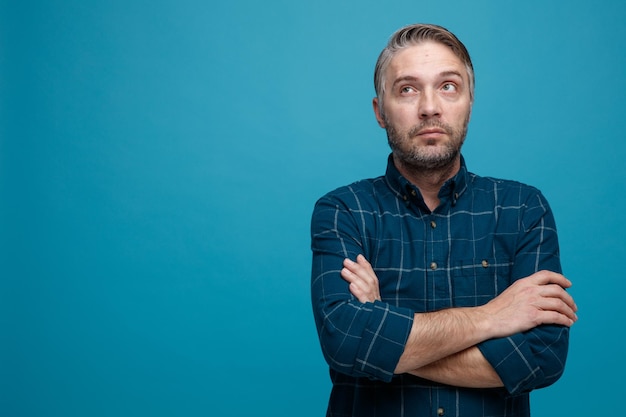 middle age man with grey hair in dark color shirt looking up with crossed hands on his chest thinking standing over blue background