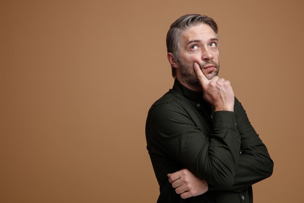 Middle age man with grey hair in dark color shirt looking up with pensive expression thinking holding hand on his chin standing over brown background