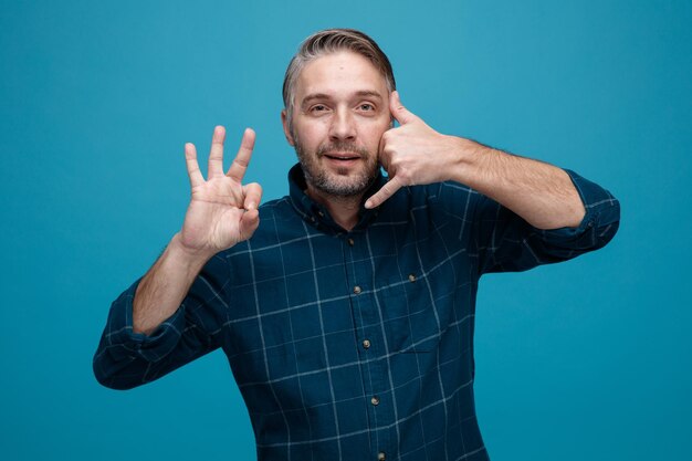 Middle age man with grey hair in dark color shirt making call me gesture showing ok sign happy and positive smiling standing over blue background