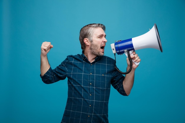 Free photo middle age man with grey hair in dark color shirt shouting in megaphone being excited and angry clenching fist standing over blue background