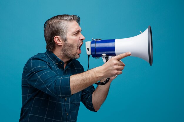 Middle age man with grey hair in dark color shirt shouting in megaphone being excited pointing with index finger at something standing over blue background