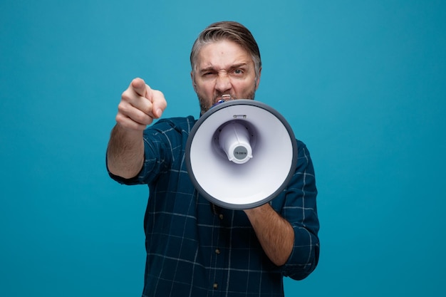 Middle age man with grey hair in dark color shirt shouting in megaphone with aggressive expression pointing with index finger at camera standing over blue background