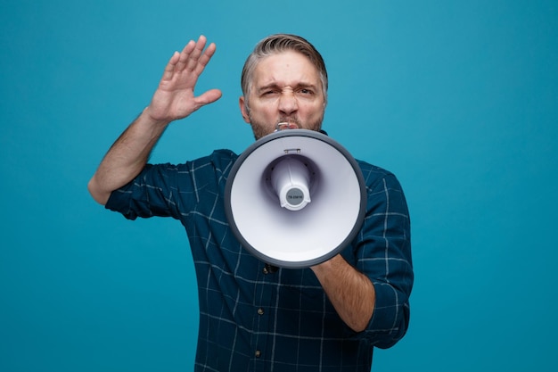 Middle age man with grey hair in dark color shirt shouting in megaphone with aggressive expression raising arm standing over blue background
