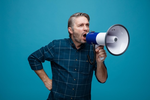 Middle age man with grey hair in dark color shirt shouting in megaphone with aggressive expression standing over blue background