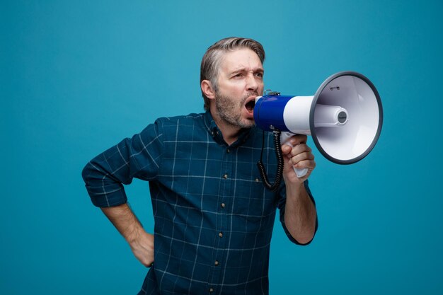 Мужчина средних лет с седыми волосами в рубашке темного цвета кричит в мегафон с агрессивным выражением лица, стоя на синем фоне