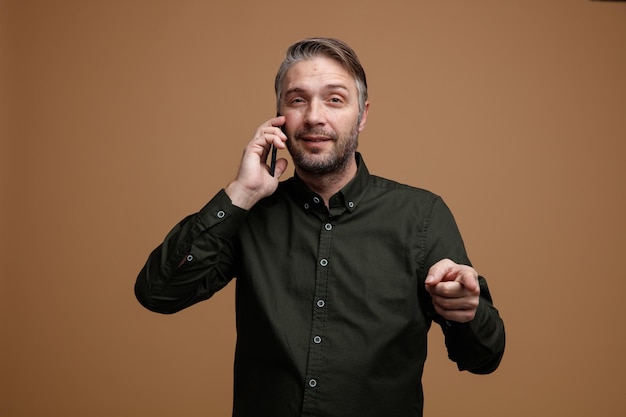 茶色の背景の上に立っているカメラに人差し指で指して笑顔で携帯電話で話している暗い色のシャツの灰色の髪を持つ中年男性