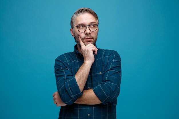 Middle age man with grey hair in dark color shirt wearing glasses looking aside with pensive expression thinking standing over blue background