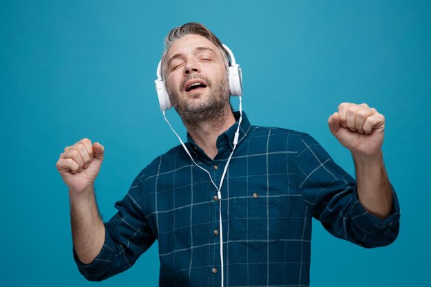 Free photo middle age man with grey hair in dark color shirt with headphones happy and pleased clenching fists listening to music relaxing standing over blue background