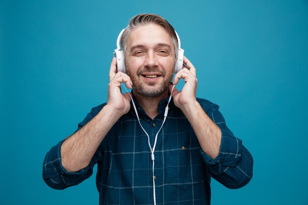 Middle age man with grey hair in dark color shirt with headphones happy and positive listening to music standing over blue background