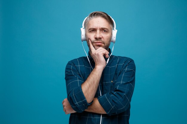 Middle age man with grey hair in dark color shirt with headphones looking aside with pensive expression being dissatisfied standing over blue background