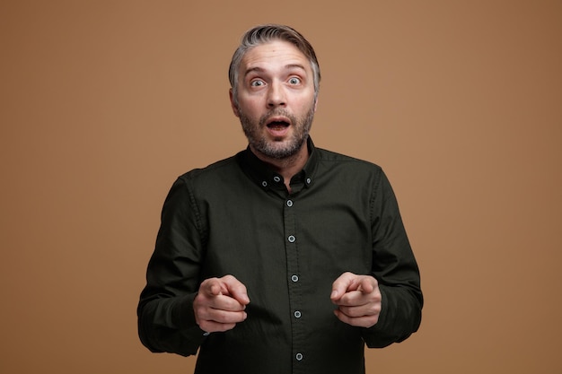 Middle age man with grey hair in dark green shirt looking at camera amazed and surprised pointing with index fingers at camera standing over brown background