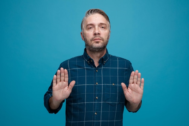 Мужчина средних лет с седыми волосами в рубашке темного цвета смотрит в камеру с серьезным лицом, делая стоп-жест руками, стоящими на синем фоне Premium Фотографии