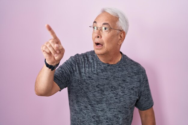 Middle age man with grey hair standing over pink background pointing with finger surprised ahead, open mouth amazed expression, something on the front