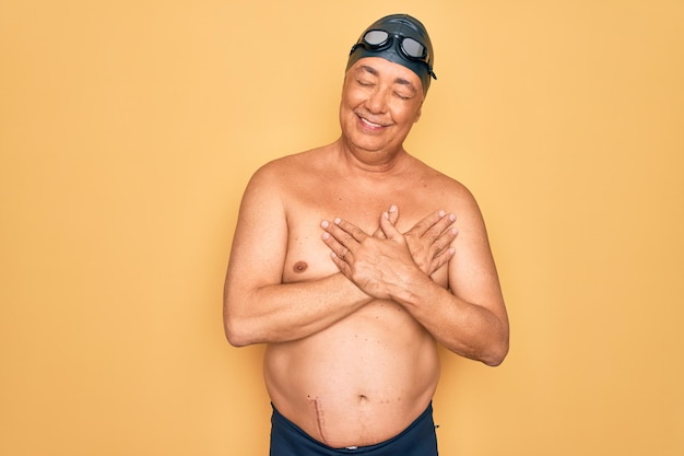 Middle age senior greyhaired swimmer man wearing swimsuit cap and goggles smiling with hands on chest with closed eyes and grateful gesture on face Health concept
