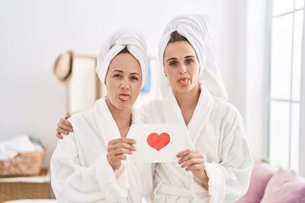Middle age woman and daughter wearing bath robe holding heart card sticking tongue out happy with funny expression.