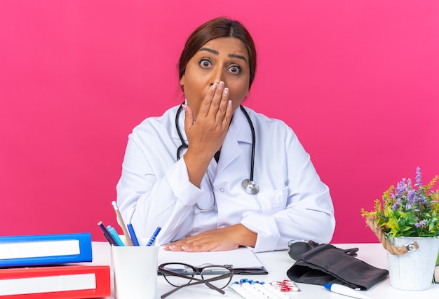 Бесплатное фото Женщина-врач среднего возраста в белом халате со стетоскопом в шоке, изогнув рот рукой, сидя за столом с офисными папками на розовом
