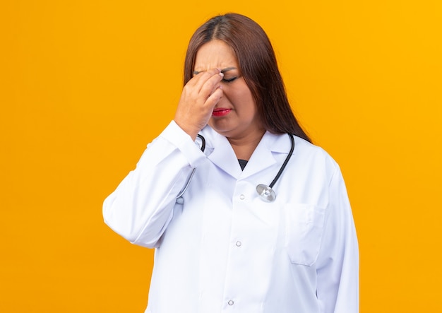 Женщина-врач среднего возраста в белом халате со стетоскопом выглядит усталой и скучающей, касаясь носа между закрытыми глазами
